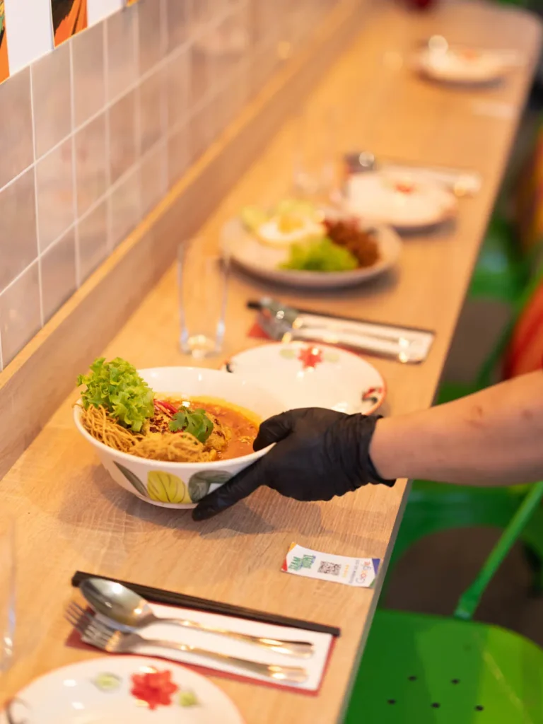 A gloved hand placing a bowl of authentic Thai Khao Soi noodle soup with crispy noodles and fresh garnish on a wooden dining counter.