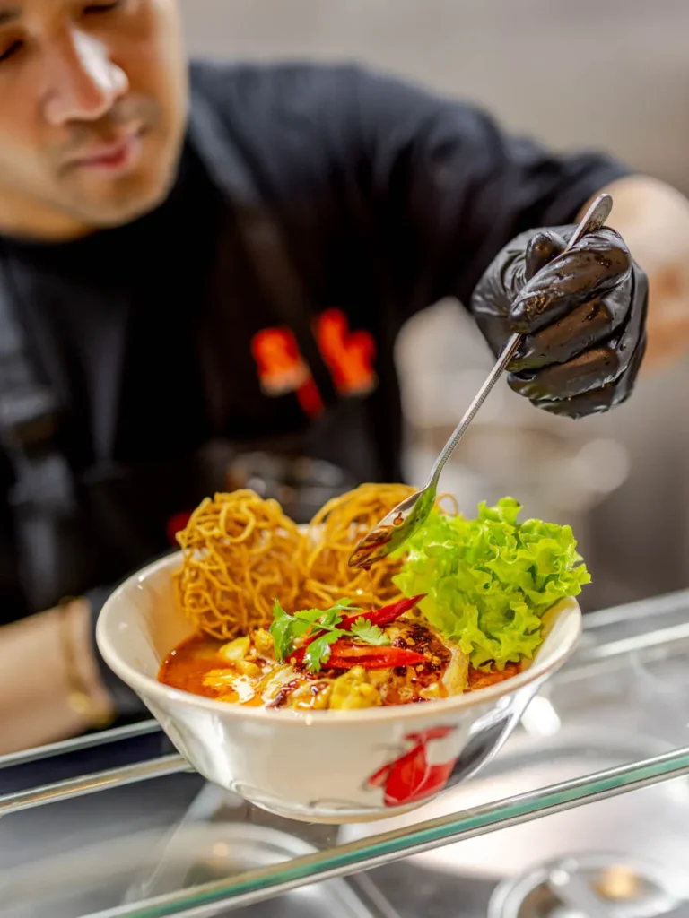 A Thai chef at Siam Bowl Amsterdam adds a final drizzle of spicy oil to a bowl of Khao Soi noodle soup with crispy noodles and fresh garnish.
