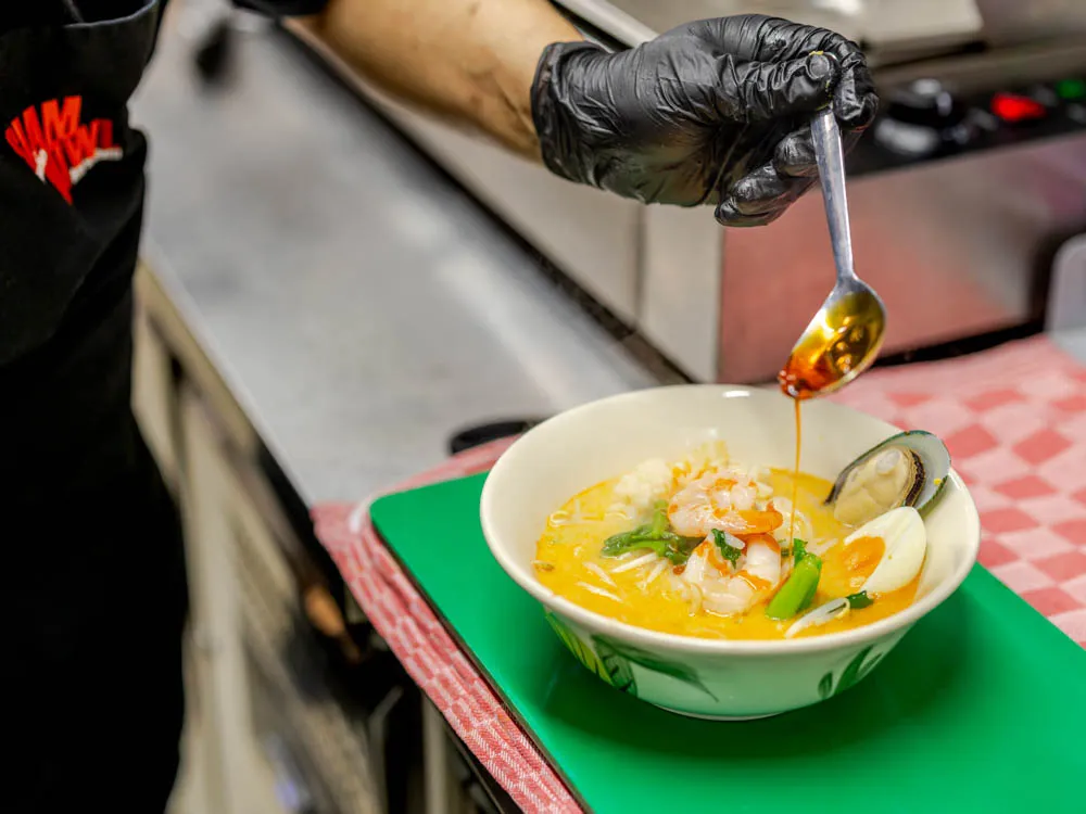 A chef at Siam Bowl Amsterdam adds spicy chili oil to a bowl of Tom Yum soup featuring fresh shrimp, mussels, and a boiled egg.