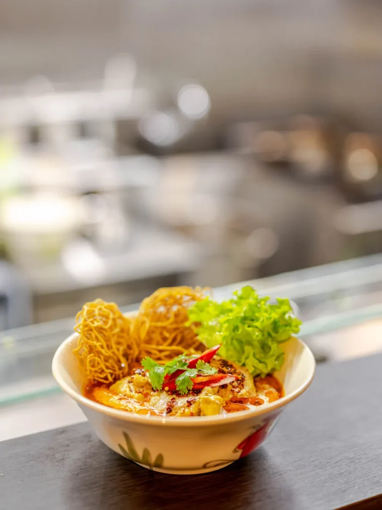 A bowl of authentic Khao Soi with crispy noodles, fresh cilantro, and red chili in a creamy curry broth at Siam Bowl.