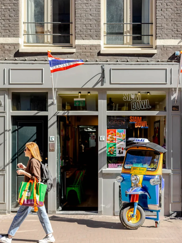 A woman walks past the Siam Bowl Thai restaurant entrance on Vijzelstraat, featuring a blue and yellow tuk tuk and neon signs.